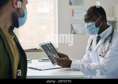 Confident mature radiologist in mask pointing at brain scan on tablet screen and looking at patient while explaining him exam results Stock Photo