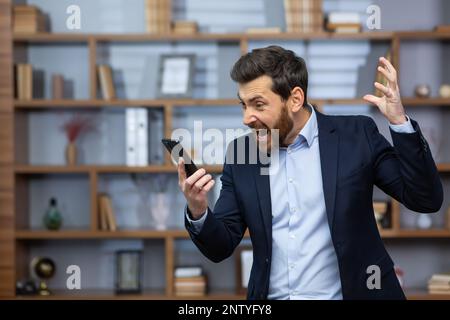 Angry boss quarreling and shouting on phone call, man in business suit at work inside office, nervously talking with colleagues using smartphone. Stock Photo