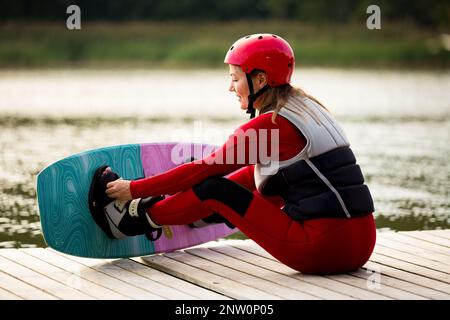 Woman in wetsuit, helmet and life vest sitting with wakeboard on a wooden pier. Sunny summer day. Safety in sport. Water sports in Finland. Insurance Stock Photo