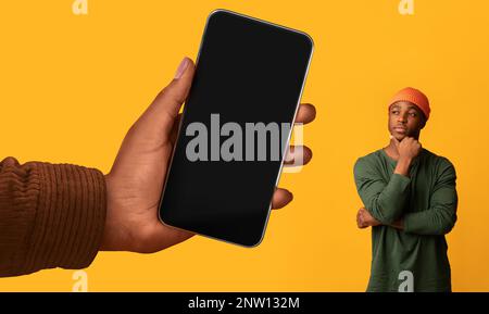 Thoughtful Young Black Guy Looking At Huge Blank Smartphone In Big Hand Stock Photo