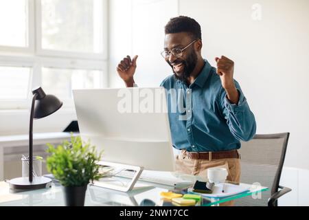 Overjoyed black businessman celebrating success with computer, looking at monitor and raising fists with excitement Stock Photo