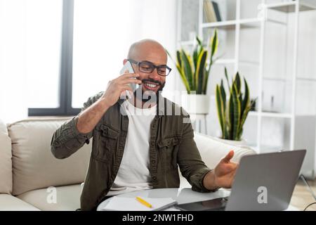 Smiling latin man using laptop and talking on cellphone, communicating with client while working online from home Stock Photo