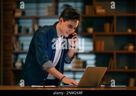Young man using laptop, talking on phone at dark office Stock Photo