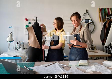 Happy young fashion designer using tablet by workplace while her mature colleague taking measures of front oart of leather attire Stock Photo