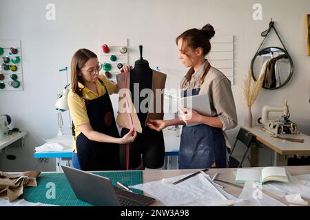 Young subordinate of tailor helping her with taking measures of unfinished leather waistcoat while both standing in front of mannequin Stock Photo