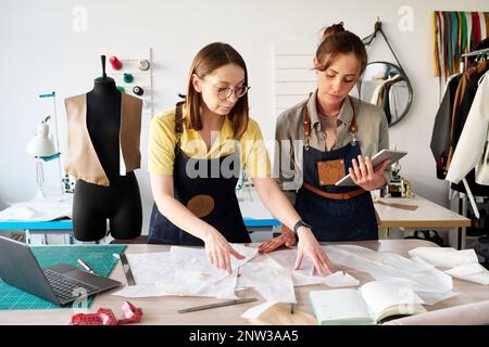 Two female tailors choosing sewing paper patterns by workplace in studio or atelier while working over new collection of leather attire Stock Photo