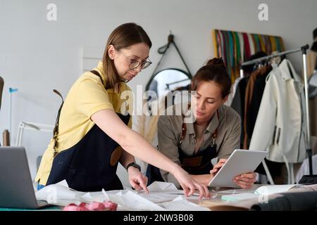 Experienced tailor and her young assistant with tablet looking at sewing paper pattern while working over creation of new attire in atelier Stock Photo