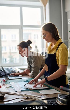 Side view of female tailors working in atelier or studio of fashion while one of them networking and the other going to circle sewing patterns Stock Photo