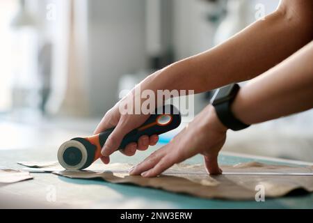 Hands of young female tanner with sharp knife cutting outline of sewing pattern out of leather piece before stitching together parts of new item Stock Photo