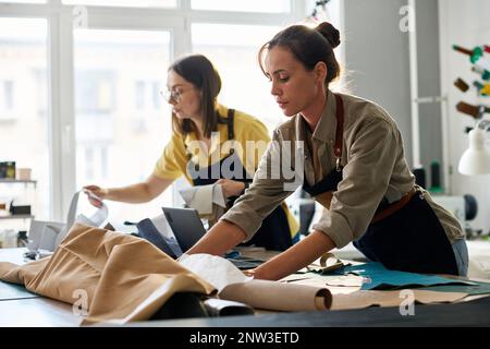 Young female tanner working with sewing patterns and leather textile while her colleague looking through new samples in atelier or studio Stock Photo