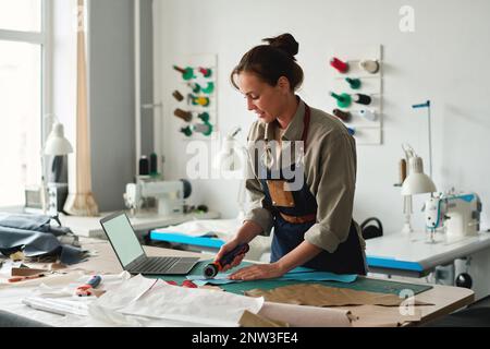 Happy young female tanner in apron bending over workplace in studio or atelier while cutting piece of blue leather with sharp knife Stock Photo