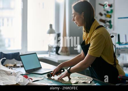 Side view of creative female fashion designer in workwear cutting piece of leather with sharp knife while bending over workplace in atelier Stock Photo