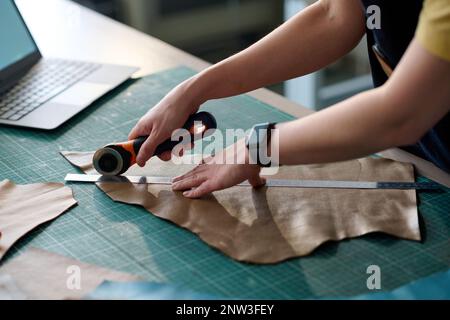 Hands of young female tailor cutting piece of beige leather on table while working over creation of new attire item in workshop or studio Stock Photo