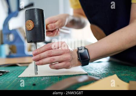 Professional female tanner keeping hammer on top of small steel shaft while pressing it down leather piece during work in craft shop Stock Photo