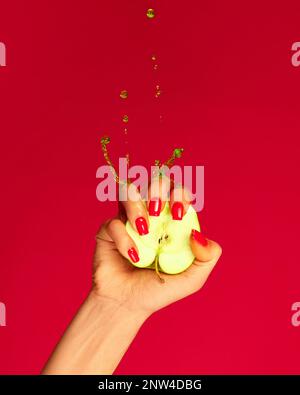 Closeup female hand with red painted nails squeezing half of apple fruit over magenta background. Contemporary art. Drops of juice fly up Stock Photo
