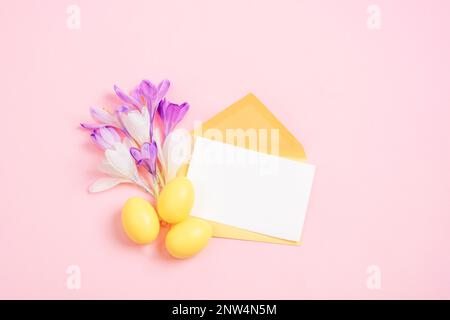 Envelope with blank card, purple crocus flowers, yellow Easter eggs on pink background. Easter greeting card. Top view, flat lay, mockup. Stock Photo