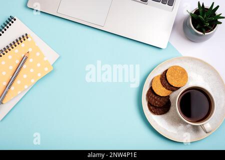 A cup of coffee with cookies, laptop computer, notepads and succulent plant on blue background. Cozy workplace, work from home concept. Top view, flat Stock Photo
