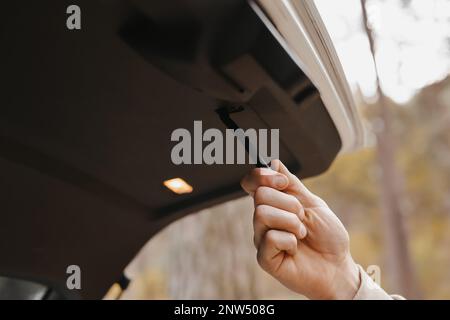 Unrecognisable man holds trunk handle and closes car trunk door outdoors in park. Interior details of prestige modern vehicle. Stock Photo