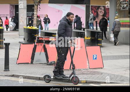 Birmingham, 28th February 2023 - A man illegally rides his personal electric scooter past an empty Voi electric hire scooter parking rack in Birmingham city centre - Voi Scooters, the only available hire scooter company in Birmingham will be shutting down their operations on Tuesday 28th February at 11pm. The electric scooters were already thin on the ground in England's Second City after the company said they would be removing the vehicles over the next few weeks. The company ran hire scooters for a trial period but have decided not to continue with the contract and no other company have step Stock Photo