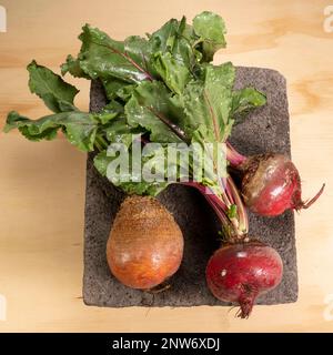 Fresh red beets with leafy beet greens on black stone plate over wooden table, viewed from above. Stock Photo