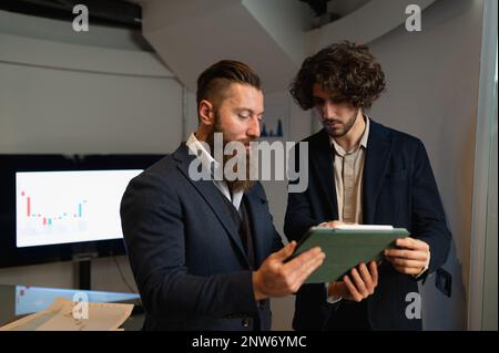Two hipster colleagues with suits discussing about work using tablet. Charts and graph in the office. Stock Photo