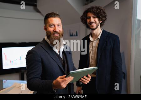 Two businessmen looking at camera posing with smiley positive expression. Colleagues wearing suits and using digital tablet. Enjoy working, new projec Stock Photo