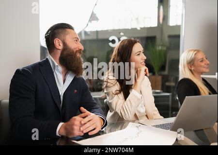 Close-up colleagues during a corporate meeting sitting at desk with paperwork and laptop listening with positive face. Stock Photo
