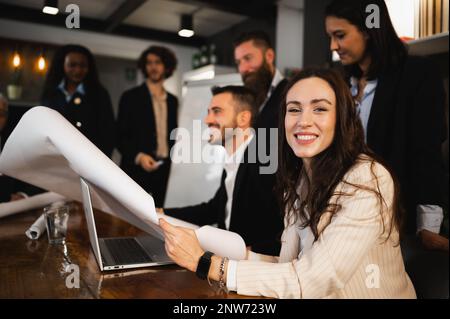 portrait of young woman smiling at camera holding a paper or blueprint. Business people having work meeting. Stock Photo