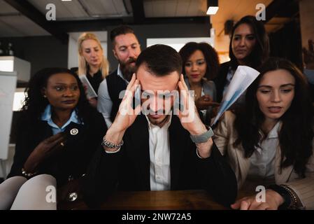 Close-up man sitting at desk with headache because colleagues are putting pressure on him. Burnout concept. Stock Photo