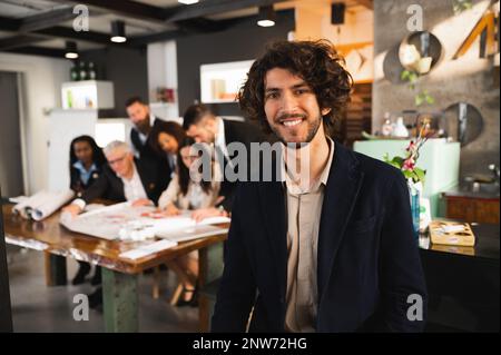 Young hipster man posing at camera smiling. Multiracial Businesspeople working at workplace in background Stock Photo