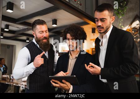 Three young businessmen discussing about something using digital tablet in workplace. Stock Photo