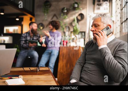 senior businessman talking on the phone sitting at desk in a modern office while colleagues discussing on background. Stock Photo