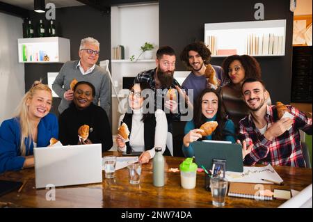 Large group of colleagues with different ethnicity sitting at desk having a break with croissants. People looking at camera smiling. Stock Photo