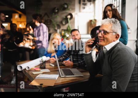 Senior man sitting at table talking on the phone. Mixed group of colleagues during a meeting Stock Photo