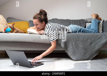 Young woman relaxing on sofa with her small dog and using laptop. Stock Photo