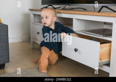 Toddler baby boy open cabinet drawer with his hand. Child explore what is in cabinet. Baby curiosity and child development stages Stock Photo