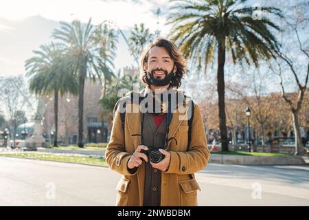 Young bearded tourist blogger standing at the avenue smiling and looking at camera. Front view of happy hipster male in a journey. Vacation concept. High quality photo Stock Photo