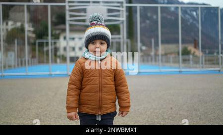 Small baby boy stands outside at park during winter season wearing beanie scarf and jacket. Pensive toddler child stands outdoors with warm clothes Stock Photo