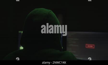 Mysterious Hacker or Programmer with Hoodie Hacking in a Desolate Room with Green Lights, Back View Shot Stock Photo