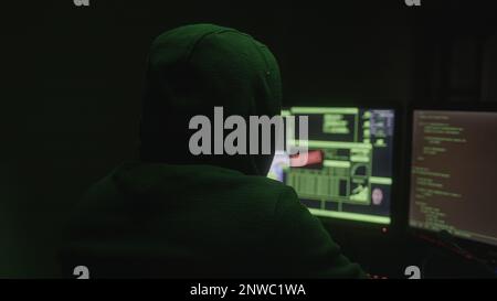 Hacker or Programmer with Hoodie Hacking in a Desolate Room with Green Lights, Back View Shot Stock Photo