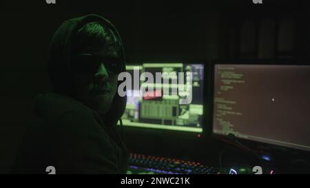 Black Hooded and Glasses-Wearing Mysterious Hacker or Programmer Hacking in an Abandoned Room with Green Lights Suddenly Turns to Look at the Camera, Stock Photo