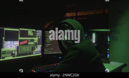 Black Glasses-Wearing Hooded Young Hacker with Two Monitors in Front Hacking and Looking at Camera, Close-up Shot Stock Photo
