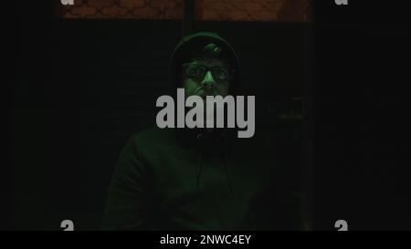The Mysterious Hooded Handsome Hacker with Glasses in a Green-Lit Abandoned Underground Room, Crosses Arms and Gazes at the Camera Stock Photo
