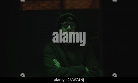 Hooded Handsome Hacker with Glasses in a Green-Lit Abandoned Underground Room, Crosses Arms and Gazes at the Camera Stock Photo