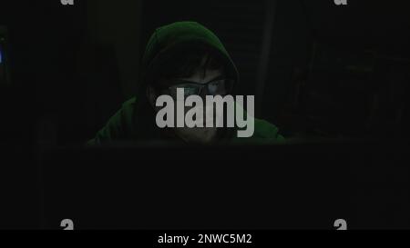 World of Hacking, Young Programmer in Hoodie and Black Glasses Immersed in Coding with Green Illumination Stock Photo