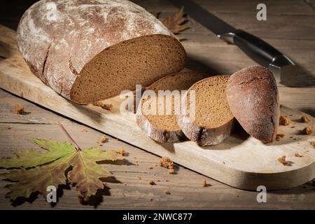 Sliced peasant black rye bread on a wooden table. In the foreground is an autumn maple leaf, in the background is a knife for cutting bread. Vignetted Stock Photo