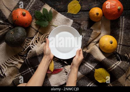 white plate in the hands of the girl on the table on a tablecloth in a cage and next to pumpkins on an old wooden table, festive serving decor, plate Stock Photo