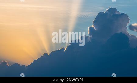 Dramatic sunset with long sunbeams on summer cloudscape at scenic evening dusk. Beautiful contrast between yellow sun beams and dark blue cloud on sky. Stock Photo