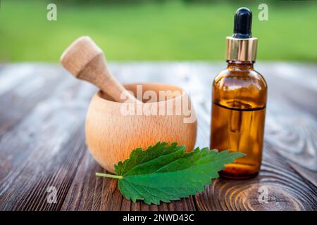Elexir and nettle essential oil. Wooden mortar and cosmetic jar with dispenser on the table near a leaf of fresh nettle Stock Photo