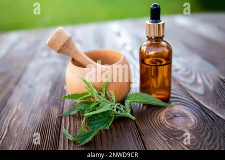 Elexir and nettle essential oil. Wooden mortar and cosmetic jar with dispenser on the table near a leaf of fresh nettle Stock Photo